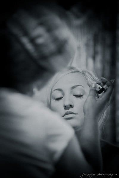 Black and white image of a person with closed eyes having makeup applied. Another person’s hands are visible, holding a makeup brush near the persons face. The background is softly blurred.