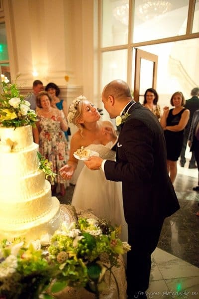 A bride and groom stand beside a tiered wedding cake. The bride leans towards the groom as they prepare to cut the cake. Guests watch in the background, smiling and capturing the moment with their cameras.
