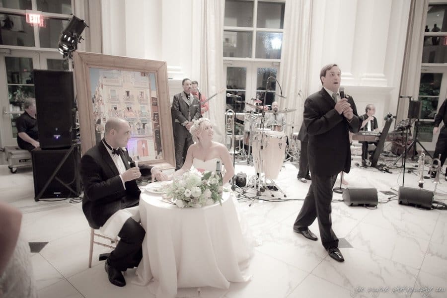 A wedding reception scene with the bride and groom seated at a table. A man in a suit stands nearby, speaking into a microphone. Musicians with instruments are in the background, and a painting is displayed on an easel.