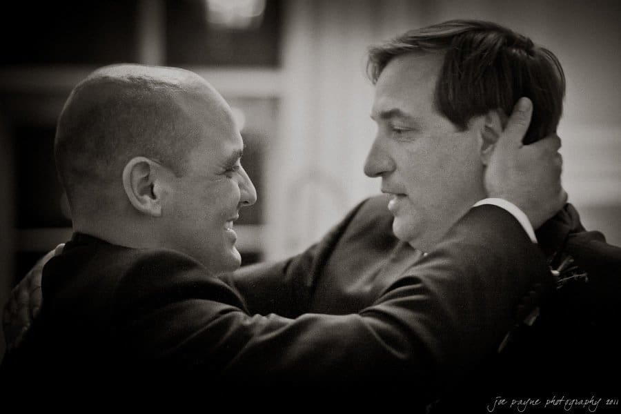 Two men exchange a warm embrace, facing each other closely. One man holds the others face with both hands. The image is in black and white, capturing a candid and emotional moment.