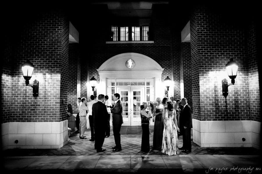 A black and white photo of a group of people dressed in formal attire standing outside a building with large brick columns and bright exterior lights. The scene appears to be an evening event or gathering.