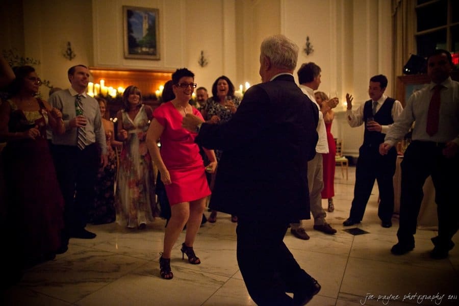 People are dancing in a softly lit room. A woman in a red dress and a man in a suit are prominently engaged in dancing, surrounded by others who are watching and enjoying the moment. The atmosphere appears lively and festive.