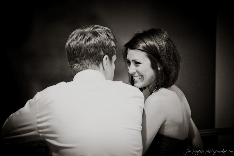 A black and white image of a couple sitting close together, with the woman smiling at the man. The mans back is to the camera, and they appear to be engaged in a happy and intimate conversation.