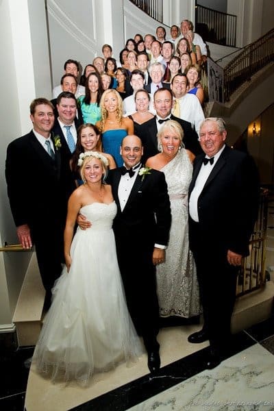 A large group of people, including a bride in a white gown and groom in a tuxedo, stand on a staircase and smile at the camera. The group is dressed in formal attire, with several women in dresses and men in suits or tuxedos.