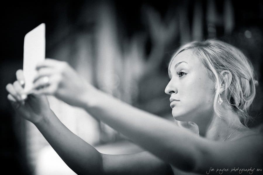 A woman with short blonde hair is taking a selfie with a smartphone, looking intently at the screen. The image is in black and white, with a blurred background.