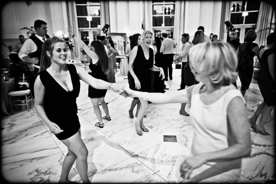 Black and white photo of a lively dance floor scene at a formal event. Several people are dancing energetically, with one couple in the foreground holding hands. The room is decorated elegantly, featuring large windows and chandeliers.