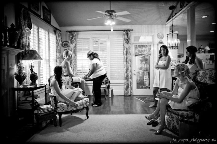 A group of women in elegant dresses gathers in a cozy, vintage-style room with patterned chairs and wooden flooring. One woman stands adjusting something, while others are seated or standing, engaged in conversation. A ceiling fan spins overhead.