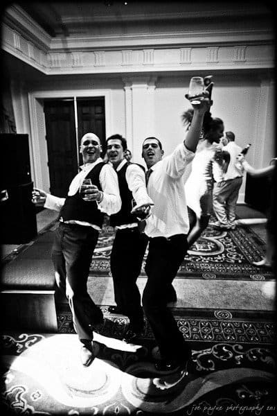 Three men in formal attire joyfully pose together on a patterned carpet, holding drinks and smiling, with one man kicking his leg high. The setting appears to be a ballroom or large indoor space, with other people in the background.