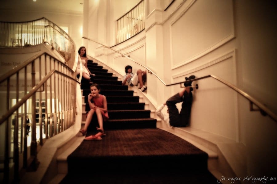 Four people are sitting on a large, carpeted staircase inside a building. The scene has a soft focus and warm lighting. Two individuals are seated casually on the steps, while two others are leaning against the wall.