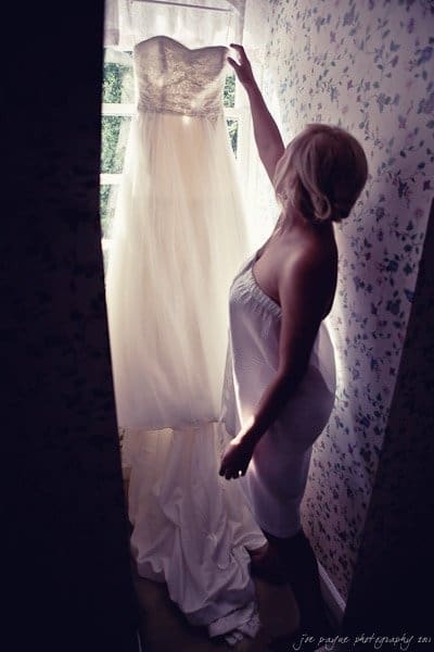 A person in a strapless dress holds up a wedding gown in front of a window with floral-patterned wallpaper. The sunlight filters through the window, highlighting the gowns details.