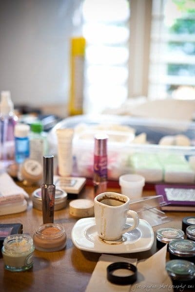 A cluttered table filled with makeup items, including foundation, mascara, and jars of cream. A cup of coffee in a saucer sits among the products. The background shows a window with blinds partially open, letting in soft light.