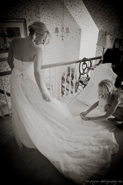 A bride in a strapless gown stands on a staircase, holding her train. Another woman kneels, adjusting the dress. The setting is a warmly lit, vintage-style room with floral wallpaper and framed artwork on the walls.