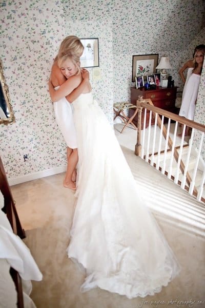 A bride in a white gown hugs a woman in a white dress on a carpeted staircase landing. The room has floral wallpaper, wooden furniture, framed photos, and a lamp. Another woman wrapped in a towel looks on from a doorway, smiling.