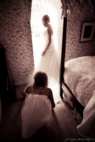 A bride in a white gown stands by a sunlit window, casting a soft glow. Another woman, also in white, kneels before her, adjusting the dress. The room has floral wallpaper and a bed with a patterned quilt.