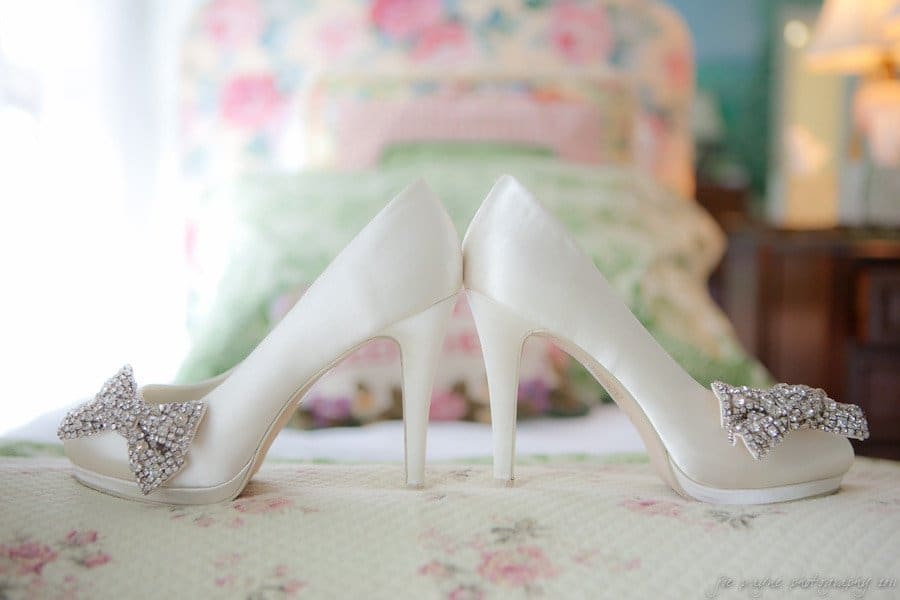 Elegant white high heels with decorative rhinestone bows, placed back-to-back on a soft quilt. The background features blurred floral patterns in pastel colors, creating a romantic and delicate atmosphere.