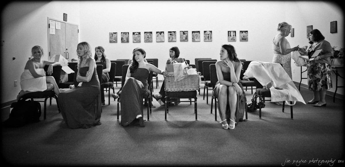 A black and white photo of a group of women in a room with chairs arranged in rows. Some are sitting, others are standing, and they seem to be preparing for an event. A table with items is visible in the background.