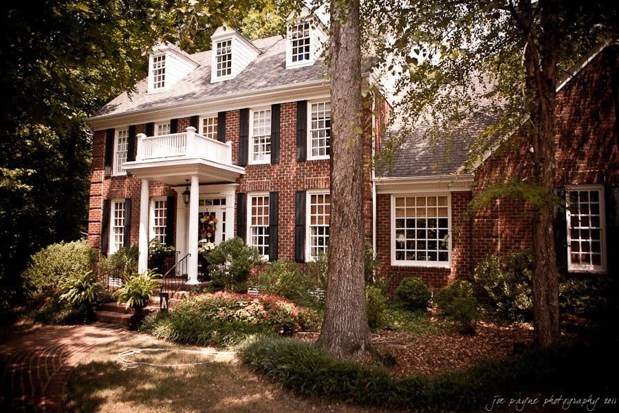 A large, two-story brick house with white trim and black shutters is surrounded by trees and greenery. A small balcony is above the entrance, and a curved brick walkway leads to the front steps.