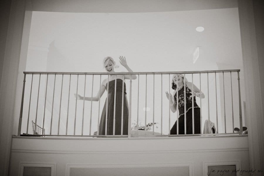 Two women joyfully wave from a balcony, each on either side of a railing. Theyre dressed in formal attire, one in a dark dress and the other in a light-colored top and dark skirt. The setting appears elegant and indoors.