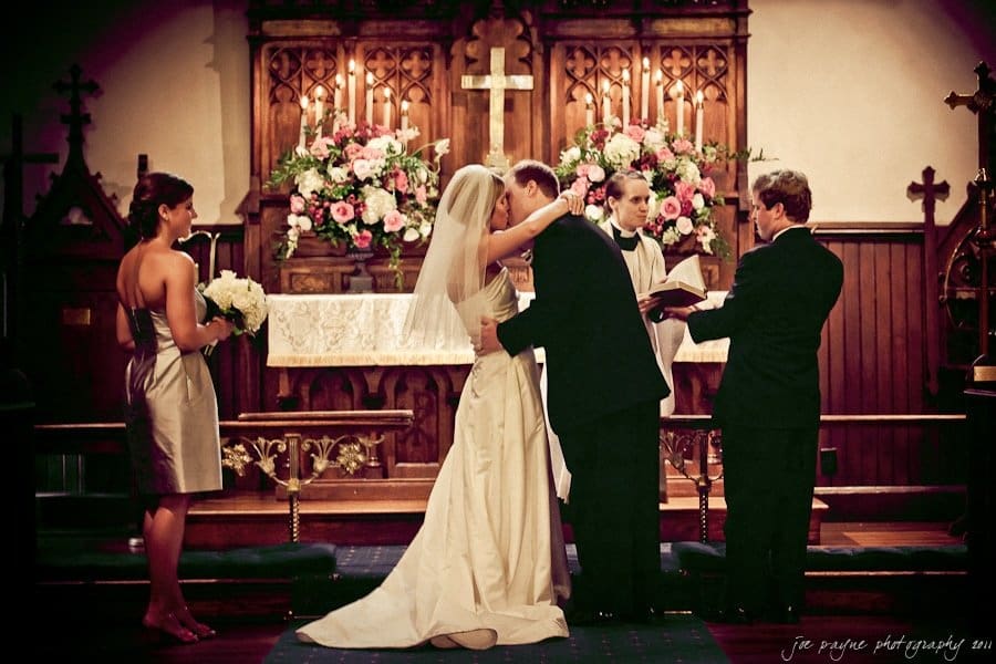 A bride and groom share a kiss at the altar during their wedding ceremony. They are surrounded by two attendants holding a bouquet and an open book. The setting is a decorated church with floral arrangements and wooden details.