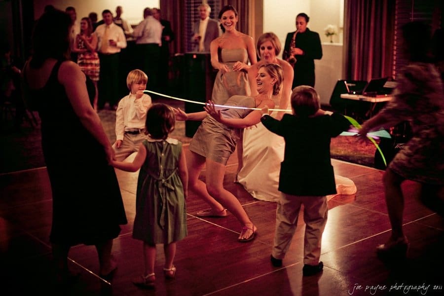 People at a lively celebration are dancing and playing limbo. Children and adults are joyfully participating in the game, with one person bending backward under a limbo stick. The atmosphere is festive and filled with laughter.