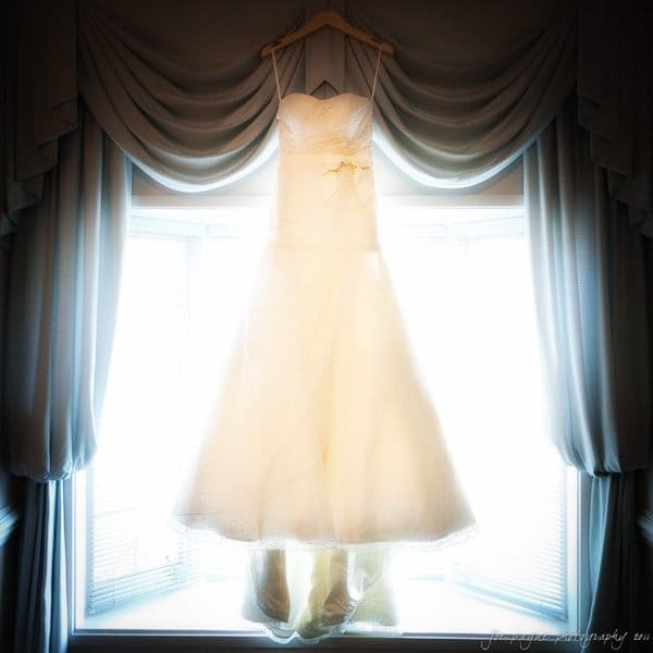 carolina inn weddings | allison and matthew A white wedding dress hangs on a wooden hanger in front of a large, bright window. The window is framed with elegant, draped curtains, creating a soft and romantic atmosphere.