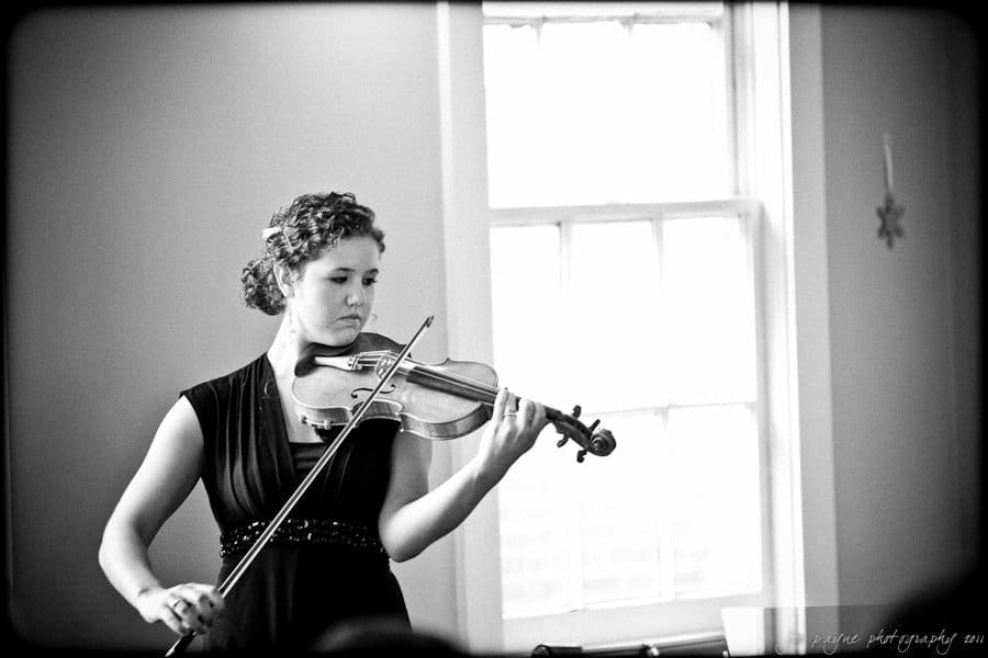 carolina inn weddings | allison and matthew A black and white image of a woman playing a violin in a softly lit room. She is wearing a sleeveless dress and focused on the music. A window in the background casts natural light into the space.