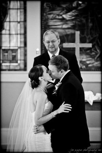 carolina inn weddings | allison and matthew A bride and groom share a kiss during a wedding ceremony. They are standing in front of a smiling officiant and a cross, with stained glass windows in the background. The couple looks happy and is dressed in wedding attire.
