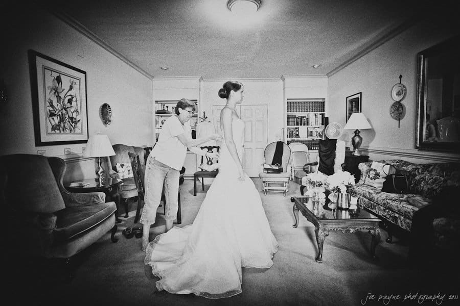 carolina inn weddings | allison and matthew A woman assists a bride with her wedding dress in a living room. The room is decorated with framed art, a floral centerpiece on a coffee table, and comfortable seating. The image is in black and white, adding a timeless feel to the scene.