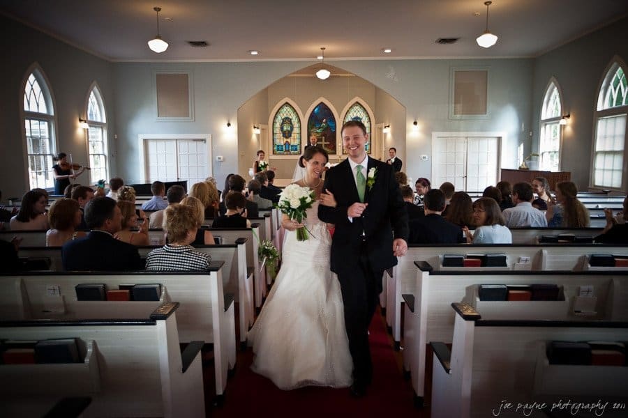 carolina inn weddings | allison and matthew A bride and groom walk down the aisle in a small church filled with guests. The bride is in a white dress holding flowers, and the groom wears a black suit with a green boutonniere. Stained glass windows and wooden pews are visible in the background.