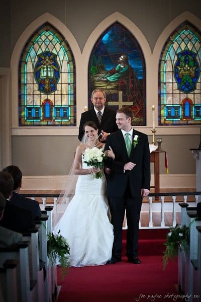 carolina inn weddings | allison and matthew A bride and groom smile as they walk down the aisle of a church, with the officiant standing behind them. The church has stained glass windows in the background, and the bride holds a bouquet of flowers.