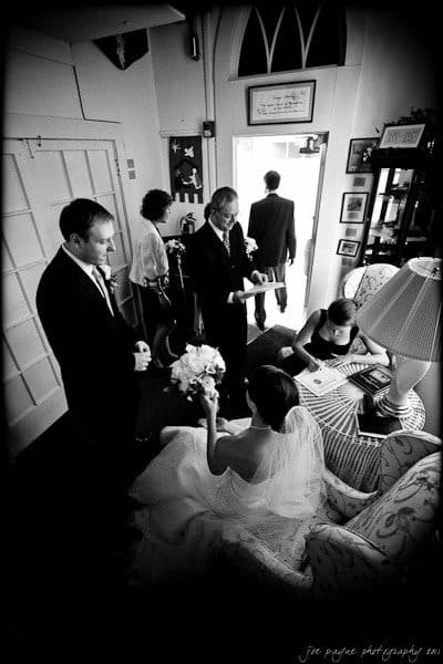 carolina inn weddings | allison and matthew A black and white photo of a bride seated on a sofa with a bouquet, surrounded by people in formal attire. A bridesmaid is writing, while others stand and chat in a cozy room. Sunlight streams through an open door, casting soft light on the scene.