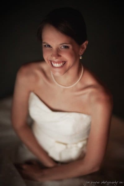 carolina inn weddings | allison and matthew A smiling bride in a strapless white wedding dress and pearl necklace looks up at the camera. The image has a soft focus, creating a warm, intimate atmosphere.