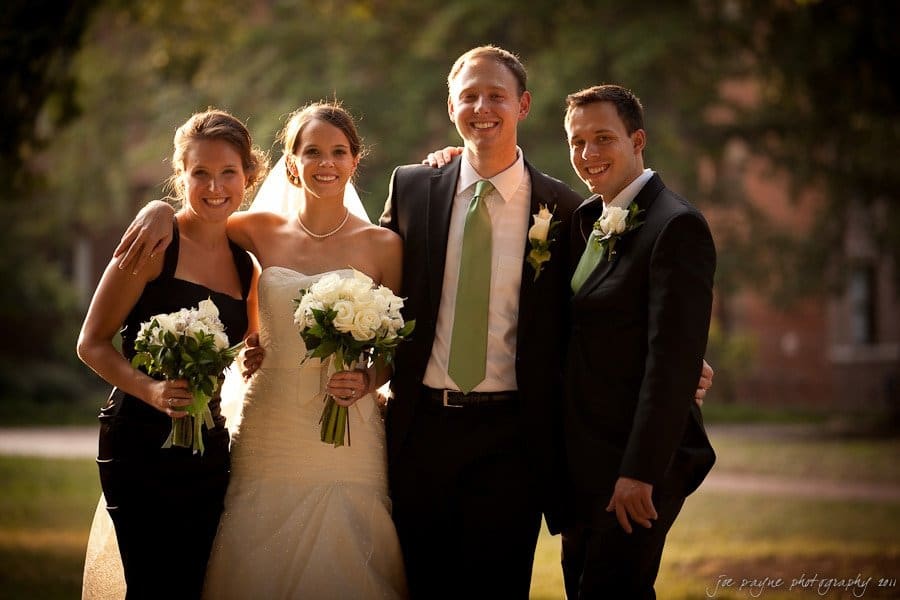 carolina inn weddings | allison and matthew A bride and groom stand arm in arm with two bridesmaids, all smiling. The bride holds a bouquet of white roses, while the bridesmaids wear black dresses. The groom and his companion both wear black suits, with the groom in a green tie.