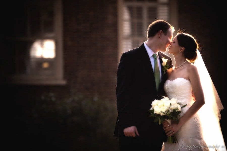 carolina inn weddings | allison and matthew A bride and groom share an intimate moment, about to kiss outdoors. The bride holds a bouquet of white roses, and both are dressed in elegant wedding attire. A brick building is softly blurred in the background, creating a romantic atmosphere.