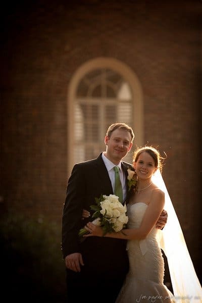 carolina inn weddings | allison and matthew A bride and groom stand together, smiling, in front of a brick building with an arched window. The bride holds a bouquet of white flowers, and the groom wears a dark suit. Soft sunlight illuminates the scene, creating a warm, romantic atmosphere.