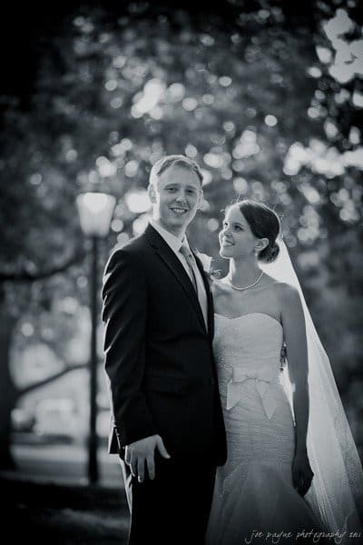 carolina inn weddings | allison and matthew A bride and groom stand closely together outdoors, smiling softly. The bride is dressed in a strapless gown with a veil, and the groom is in a dark suit. Trees and a streetlamp are blurred in the background, creating a serene atmosphere.