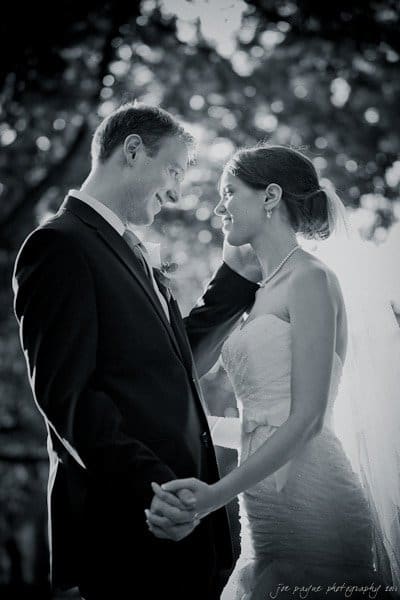 carolina inn weddings | allison and matthew A bride and groom gaze lovingly at each other, holding hands in an outdoor setting. The black-and-white photo captures them smiling, with the bride touching the grooms face. Sunlight filters through the trees, highlighting their happiness.