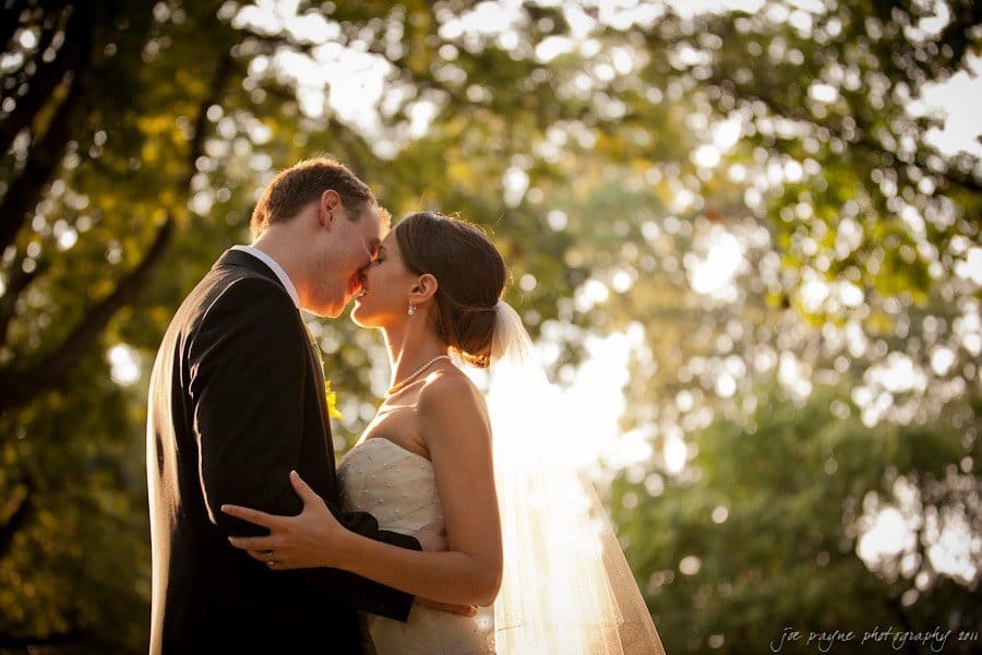 carolina inn weddings | allison and matthew A couple embracing and kissing in a sunlit, tree-filled setting. The bride is wearing a strapless gown and veil, while the groom is in a suit. Sunlight filters through the leaves, creating a warm, romantic atmosphere.