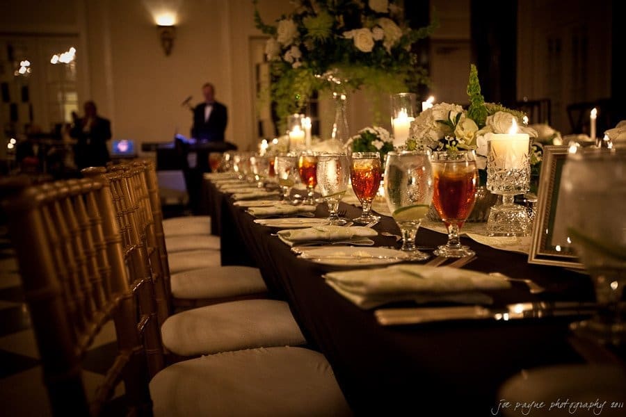 carolina inn weddings | allison and matthew A dimly lit, elegant banquet setup with a long table adorned with white floral arrangements, candles, and glasses of iced tea. The focus is on the table, while a musician performs in the background.