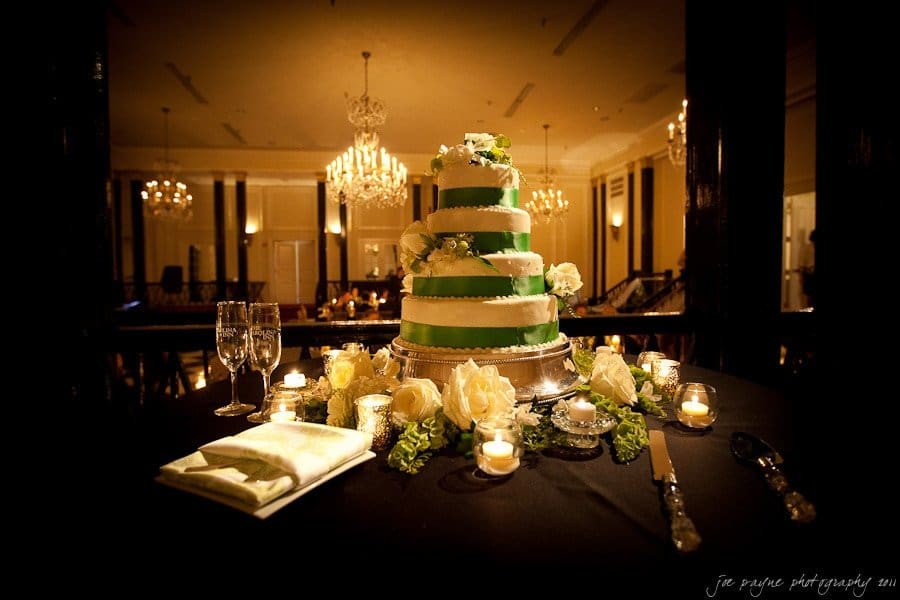 carolina inn weddings | allison and matthew A tiered wedding cake with green and white frosting is decorated with flowers on a round table. The table also has candles, roses, empty champagne flutes, and cutlery. A softly lit, elegant chandelier hangs in the background of the dimly lit room.