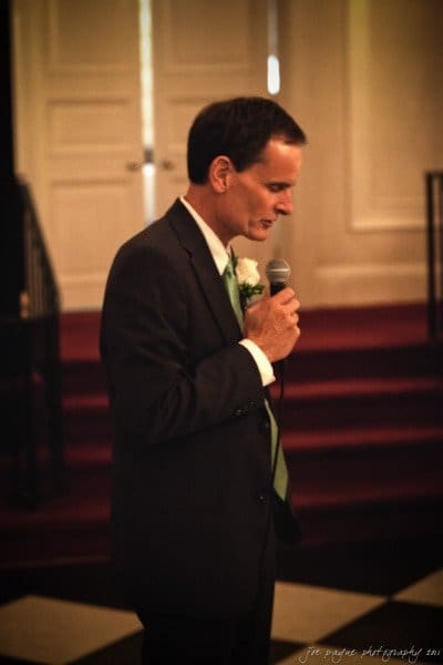 carolina inn weddings | allison and matthew A man in a dark suit and green tie is holding a microphone, standing in a dimly lit room with red carpeted steps and white doors in the background.