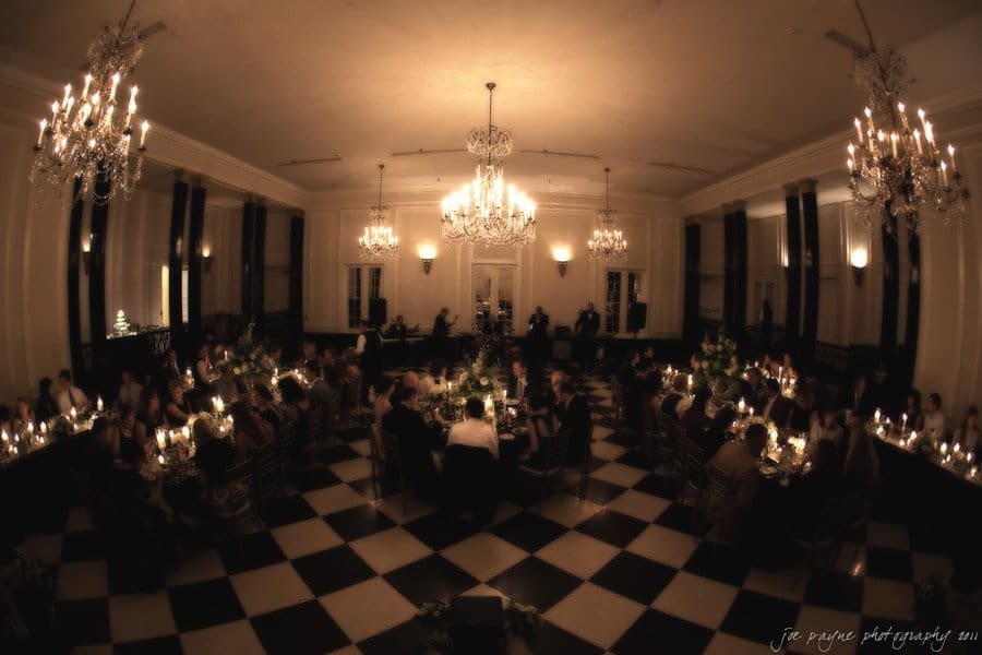 carolina inn weddings | allison and matthew A dimly lit banquet hall with chandeliers, candlelit tables, and guests seated on a checkered floor. The room has large windows and a high ceiling, creating an elegant and intimate atmosphere.