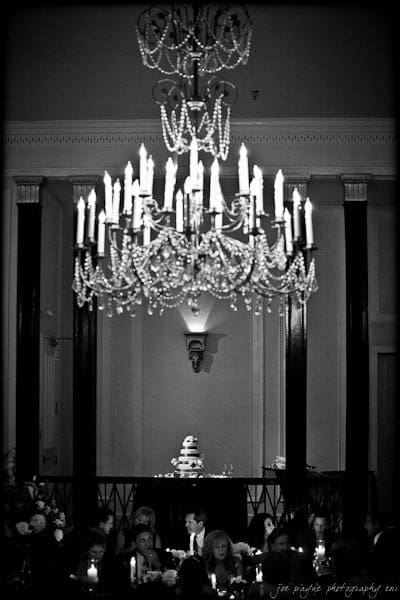 carolina inn weddings | allison and matthew A black and white photo of a grand chandelier illuminating a wedding reception. Below, guests are seated at a long table adorned with candles, and a multi-tiered cake is visible in the background.