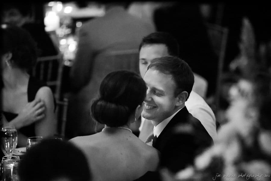 carolina inn weddings | allison and matthew A black and white photo of a couple sitting at a formal event. The man smiles, looking at the woman who is turned away from the camera. The background is softly blurred with other attendees and dim lighting.