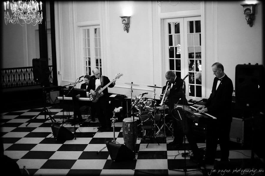 carolina inn weddings | allison and matthew A black and white photo of a jazz band performing in a formal setting. Three musicians play bass guitar, saxophone, and keyboard, with a drummer in the background. The room features a checkered floor, chandelier, and elegant decor.