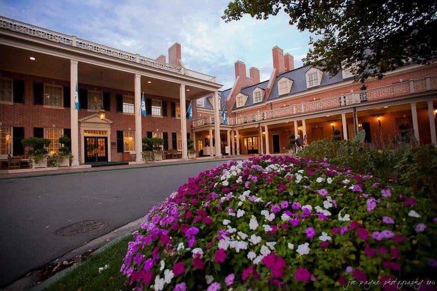 carolina inn weddings | allison and matthew A grand brick building with columns and a covered entrance is depicted, with lush, colorful flowers in the foreground. The sky is partly cloudy, and the overall scene is peaceful and elegant.