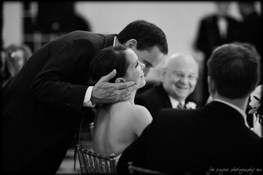 carolina inn weddings | allison and matthew A black and white photo shows a man leaning down to kiss a smiling woman seated at a table during an event. Other people are around, with one elderly man smiling and wearing glasses. The setting appears to be a formal gathering.