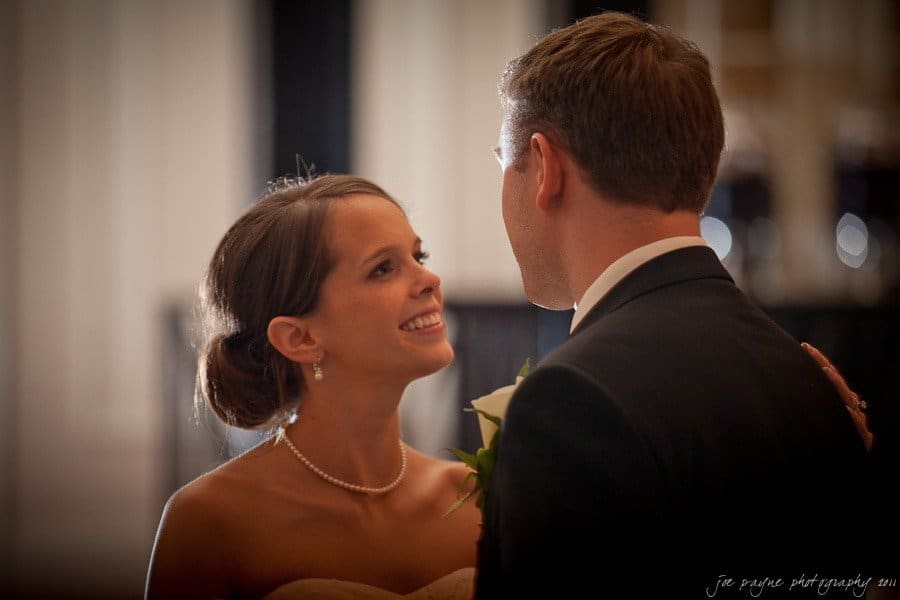 carolina inn weddings | allison and matthew A couple dressed in formal attire, with the woman smiling at the man who is holding a white flower. They are indoors, with blurred background lights creating a warm ambiance.