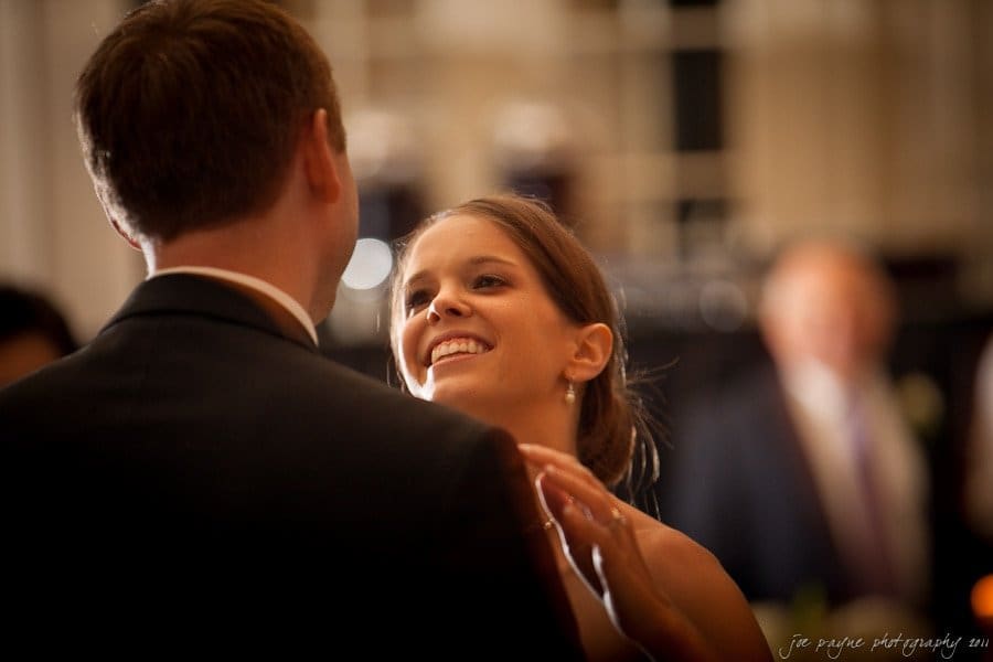 carolina inn weddings | allison and matthew A couple is dancing at an indoor event. The woman is smiling and looking up at the man, who has his back to the camera. The background is softly blurred, with another person visible in the distance.
