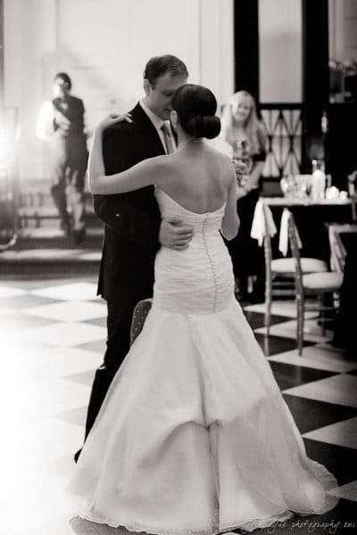 carolina inn weddings | allison and matthew A bride and groom share a first dance at their wedding. The bride is wearing a white gown, and the groom is in a dark suit. The floor is checkered, and there are a few guests in the background. The lighting is soft and romantic.